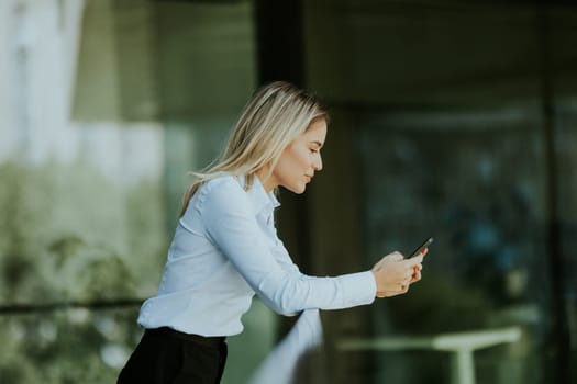 A focused woman in a white blouse engrossed in her smartphone outside a sleek modern office building on a sunny afternoon.