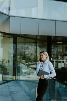 Professional businesswoman stands outside an office building, smiling as she talks on her phone while holding a laptop