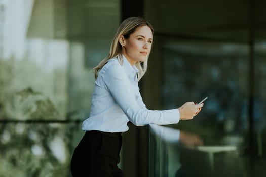 A focused woman in a white blouse engrossed in her smartphone outside a sleek modern office building on a sunny afternoon.