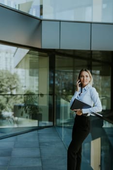 A young businesswoman smiles confidently while talking on her phone inside a sleek, modern office building, with cityscape reflections in the glass.