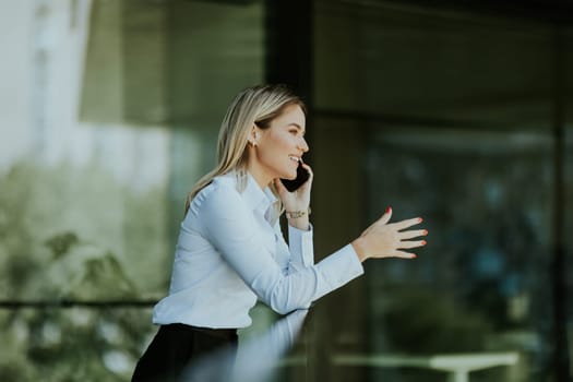 Businesswoman smiles while talking on her smartphone outside a modern office building on a sunny day, dressed in a white blouse and dark skirt