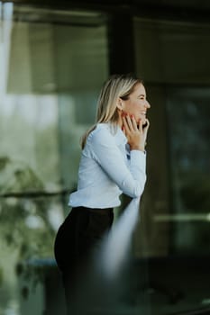 Businesswoman smiles while talking on her smartphone outside a modern office building on a sunny day, dressed in a white blouse and dark skirt