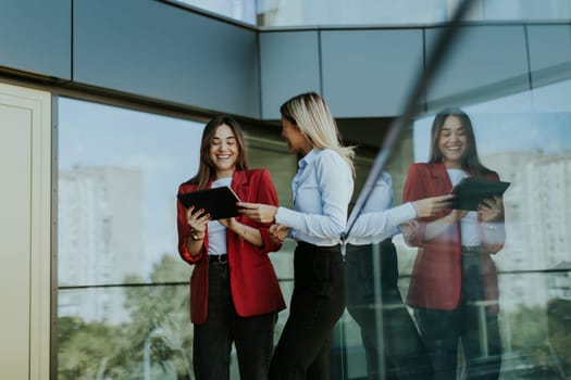 Two professional women stand outside an office building, engaging in a lively discussion while using digital tablets. Their reflections are visible on the glass facade