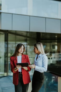 Two professional women engage in a thoughtful discussion while reviewing a tablet outside a sleek, contemporary office building during the daytime.