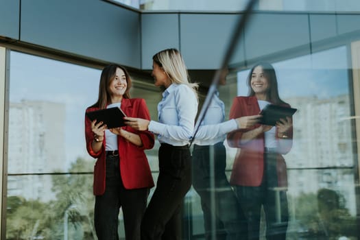 Two professional women stand outside an office building, engaging in a lively discussion while using digital tablets. Their reflections are visible on the glass facade
