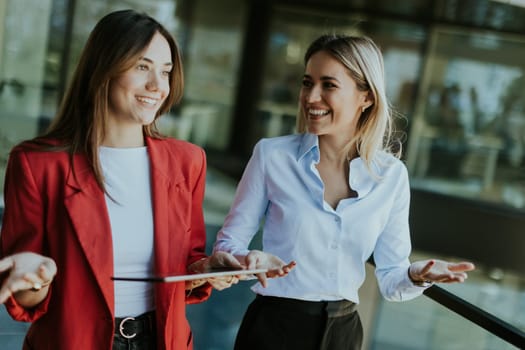 Two professional women engage in a thoughtful discussion while reviewing a tablet outside a sleek, contemporary office building during the daytime.