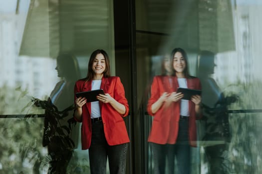 A smiling professional woman stands near a modern glass office building, holding a tablet and looking confident during the evening.