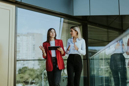Two professional women engage in a thoughtful discussion while reviewing a tablet outside a sleek, contemporary office building during the daytime.