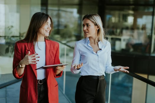 Two professional women engage in a thoughtful discussion while reviewing a tablet outside a sleek, contemporary office building during the daytime.