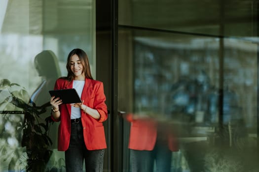 A smiling professional woman stands near a modern glass office building, holding a tablet and looking confident during the evening.