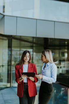 Two professional women engage in a thoughtful discussion while reviewing a tablet outside a sleek, contemporary office building during the daytime.