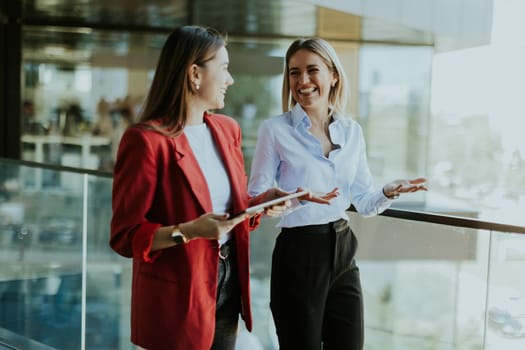 Two professional women engage in a thoughtful discussion while reviewing a tablet outside a sleek, contemporary office building during the daytime.