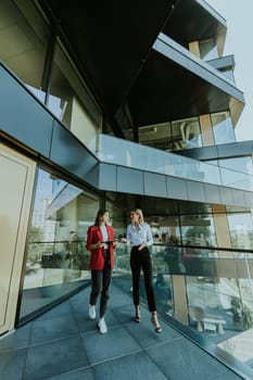 Two professional women engage in a thoughtful discussion while reviewing a tablet outside a sleek, contemporary office building during the daytime.