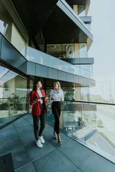 Two professional women engage in a thoughtful discussion while reviewing a tablet outside a sleek, contemporary office building during the daytime.