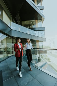 Two professional women engage in a thoughtful discussion while reviewing a tablet outside a sleek, contemporary office building during the daytime.