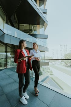 Two professional women engage in a thoughtful discussion while reviewing a tablet outside a sleek, contemporary office building during the daytime.