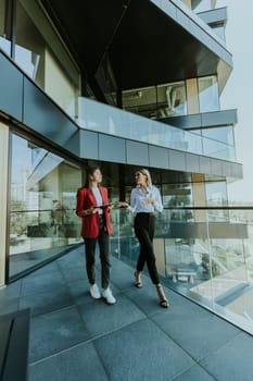 Two professional women engage in a thoughtful discussion while reviewing a tablet outside a sleek, contemporary office building during the daytime.