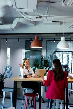 Two women engage in a discussion within a contemporary office setting, complete with trendy lighting and large windows, creating a bright and productive atmosphere.