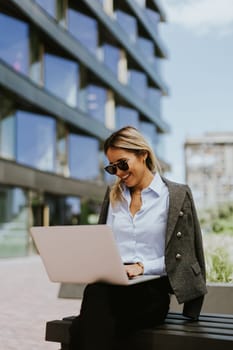 A confident businesswoman in sunglasses and a blazer works on her laptop while sitting on a bench outside a sleek, contemporary office building during a sunny day.