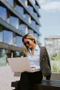 Stylish businesswoman sits on a bench outdoors, multitasking with her laptop and smartphone, enjoying the sunny weather in a contemporary city setting