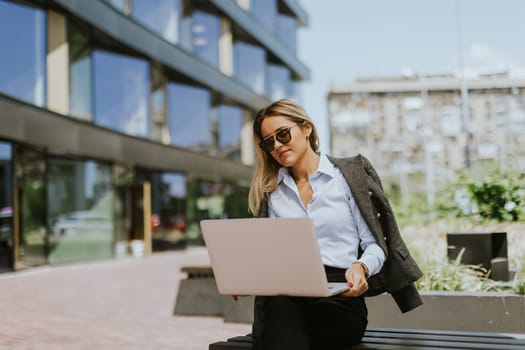 A confident businesswoman in sunglasses and a blazer works on her laptop while sitting on a bench outside a sleek, contemporary office building during a sunny day.