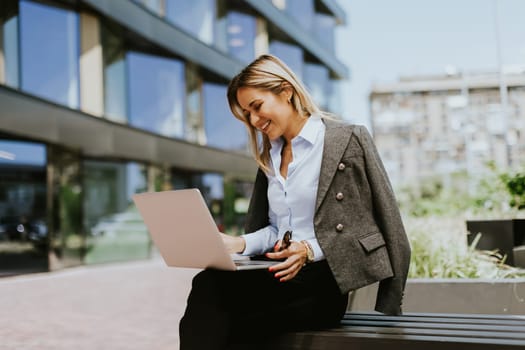 A confident businesswoman in sunglasses and a blazer works on her laptop while sitting on a bench outside a sleek, contemporary office building during a sunny day.