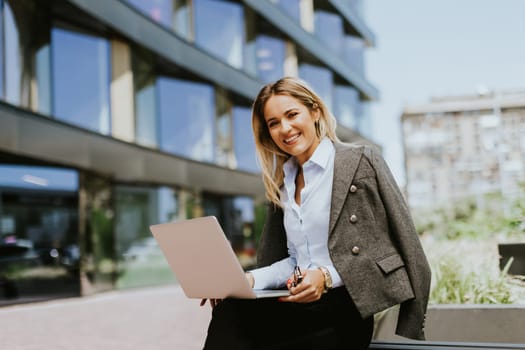 A confident businesswoman in sunglasses and a blazer works on her laptop while sitting on a bench outside a sleek, contemporary office building during a sunny day.