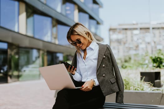 Stylish businesswoman sits on a bench outdoors, multitasking with her laptop and smartphone, enjoying the sunny weather in a contemporary city setting