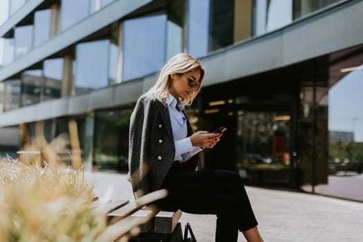 A woman with long dark hair sits on a bench, engrossed in her phone screen, her expression thoughtful and absorbed.