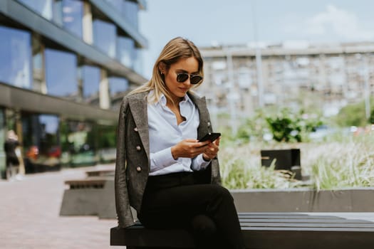 A woman with long dark hair sits on a bench, engrossed in her phone screen, her expression thoughtful and absorbed.