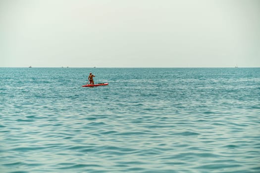 A person paddles on a red board across tranquil blue waters under a hazy sky. Nearby, small boats are visible in the distance, creating a serene atmosphere of relaxation.