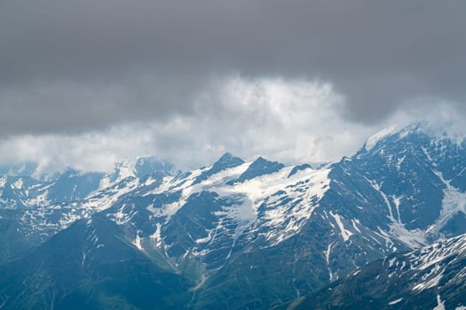 Snow-covered peaks rise dramatically against a cloudy sky. The landscape showcases rugged beauty, with patches of green highlighting the mountain slopes in midday light.