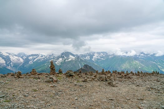 A series of stone cairns are arranged on rocky terrain, with towering snow-capped mountains and dramatic clouds in the distance. The landscape showcases the beauty of nature's formations.