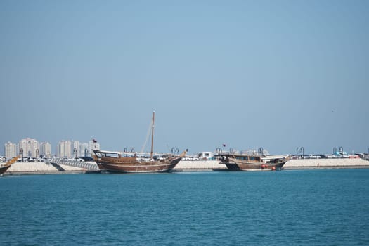 Wooden boats are anchored in a sunny harbor with tall buildings.