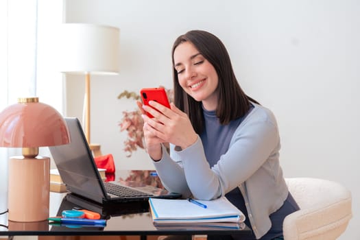 Caucasian student studying at home, using smartphone while working on a laptop. Modern workspace with books, stationery, and decor. Online learning, education, productivity, and concentration.