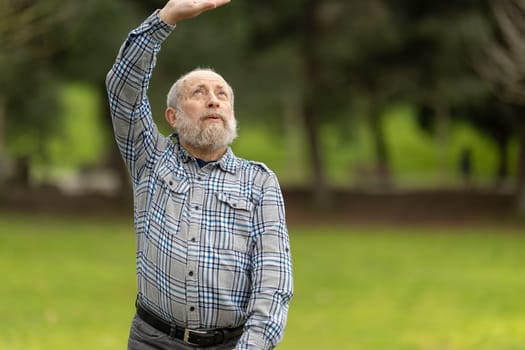 Bearded senior man shielding his eyes from the sun while stretching his arm in a park