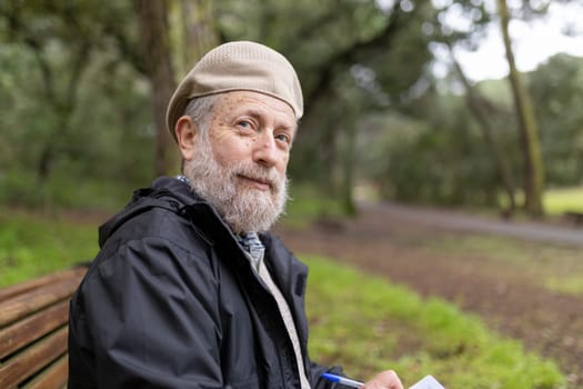 Bearded senior writer taking notes in notebook while sitting on park bench