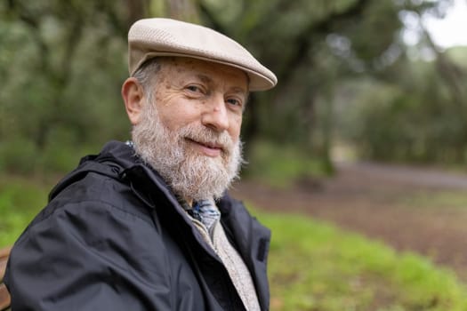 Portrait of a serene senior man with a white beard wearing a flat cap, sitting in a park and enjoying the peaceful atmosphere