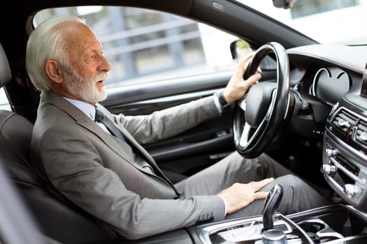An elderly gentleman, dressed in a suit, confidently drives a modern car through a bustling city street on a bright, sunny day