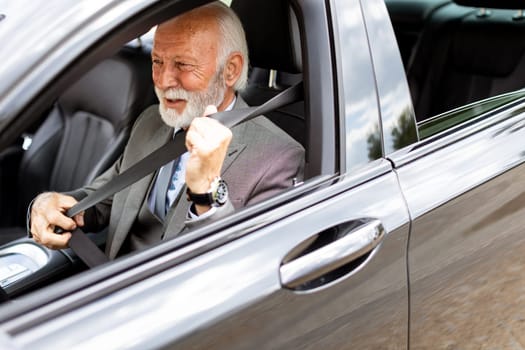 Elderly man with white hair and a beard is sitting in a modern car, fastening his seatbelt and smiling on a sunny afternoon drive
