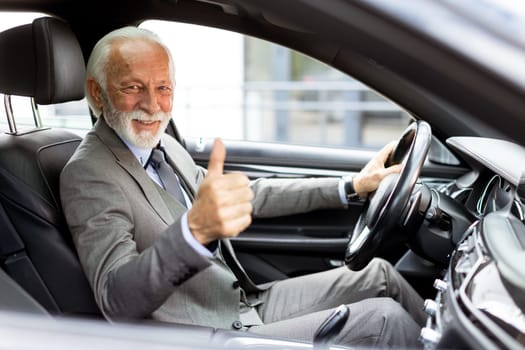 Bearded elderly man with a bright smile gives a thumbs-up while confidently driving his car through the city
