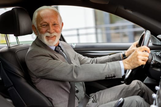 An elderly gentleman, dressed in a suit, confidently drives a modern car through a bustling city street on a bright, sunny day