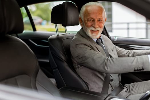 An elderly gentleman, dressed in a suit, confidently drives a modern car through a bustling city street on a bright, sunny day