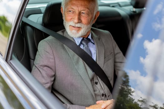 An elderly man with a beard wearing a suit and tie, secured with a seatbelt, looking content inside his vehicle