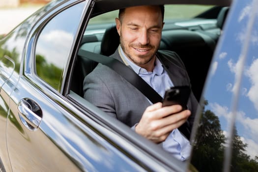 Smiling man in business attire checks his phone while seated in the backseat of a moving car