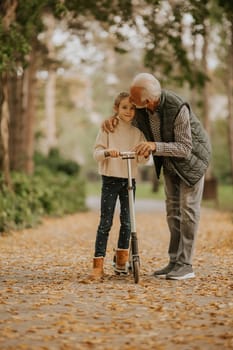 Senior man teaching his granddaughter how to ride kick scooter in the autumn park