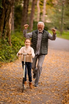 Senior man teaching his granddaughter how to ride kick scooter in the autumn park