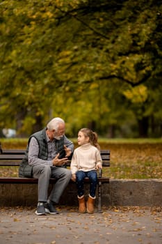 Handsome grandfather spending time with his granddaughter on bench in park on autumn day