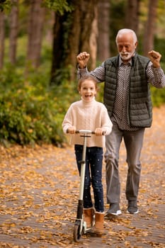 Senior man teaching his granddaughter how to ride kick scooter in the autumn park