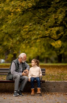 Handsome grandfather spending time with his granddaughter on bench in park on autumn day